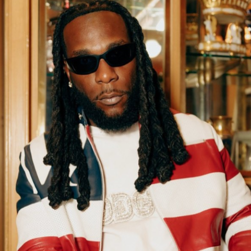 Picture of Burna Boy sitting facing the camera and spotting a white red and blue jacket. his wearing dark shades.