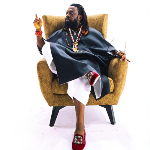 Picture of Timaya sitting on a brown plush chair and wearing what seems like a caftann and white pants